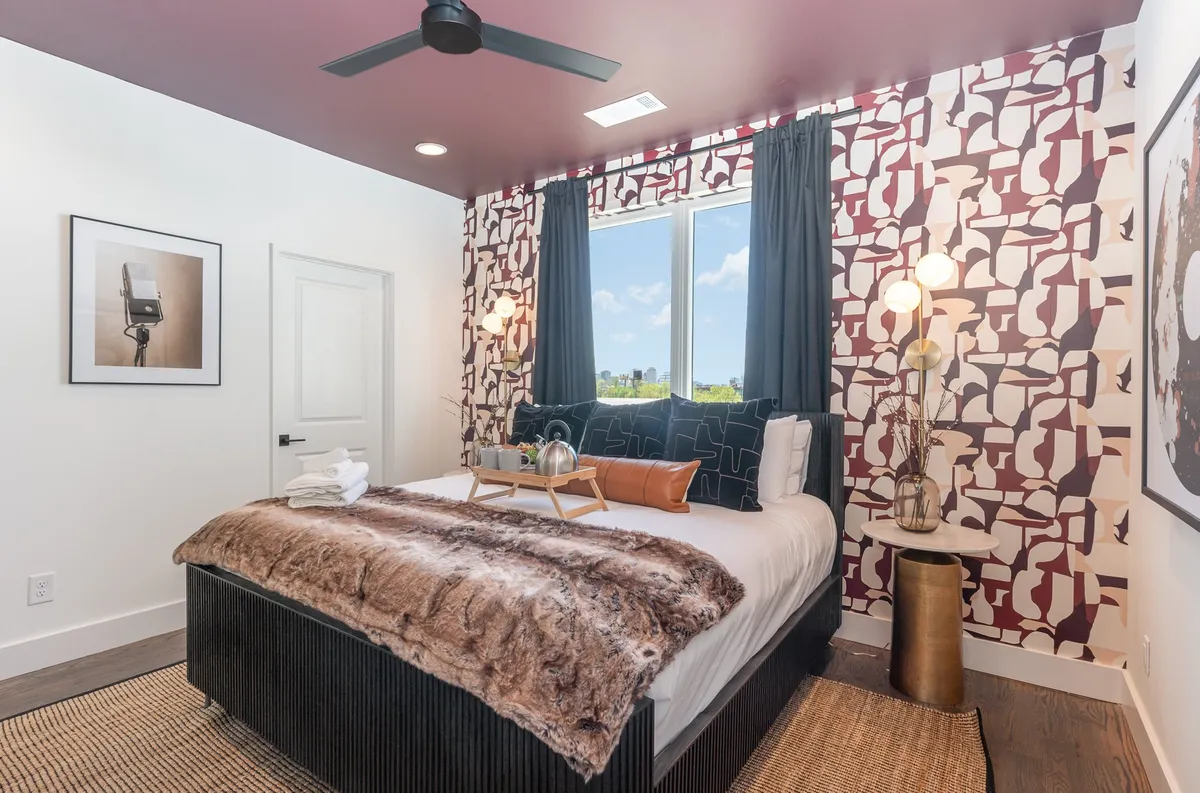 A king bedroom featuring a plush bed with layered bedding, a decorative wall with an abstract pattern, large window streaming in natural light, and stylish bedside lamps. The atmosphere is comfortable, contemporary, and inviting, ideal for relaxation.