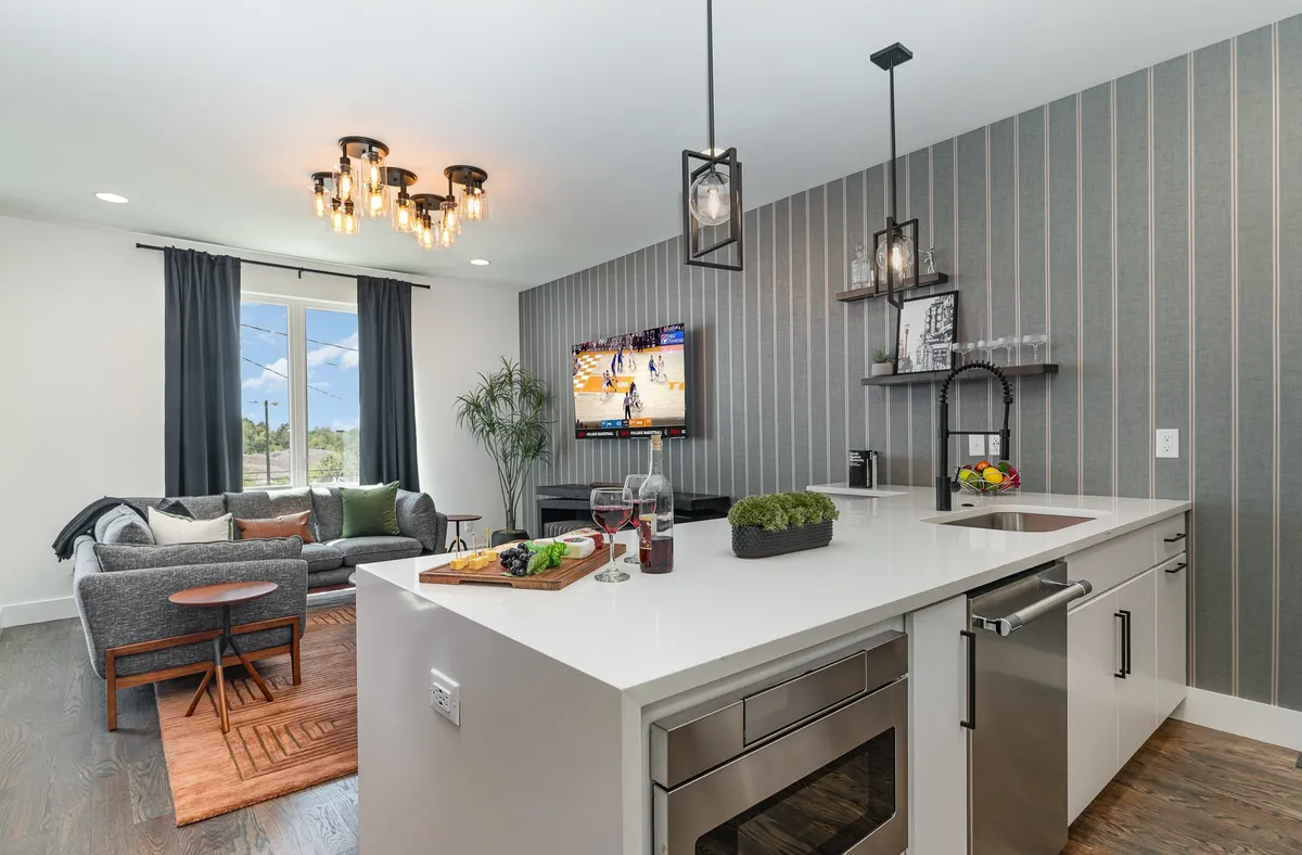 An open-plan space blending a modern kitchen island with bar seating and a comfortable seating area. The room features a television displaying a sports game, decorative striped walls, and lush plant accents for a casual, sociable vibe.