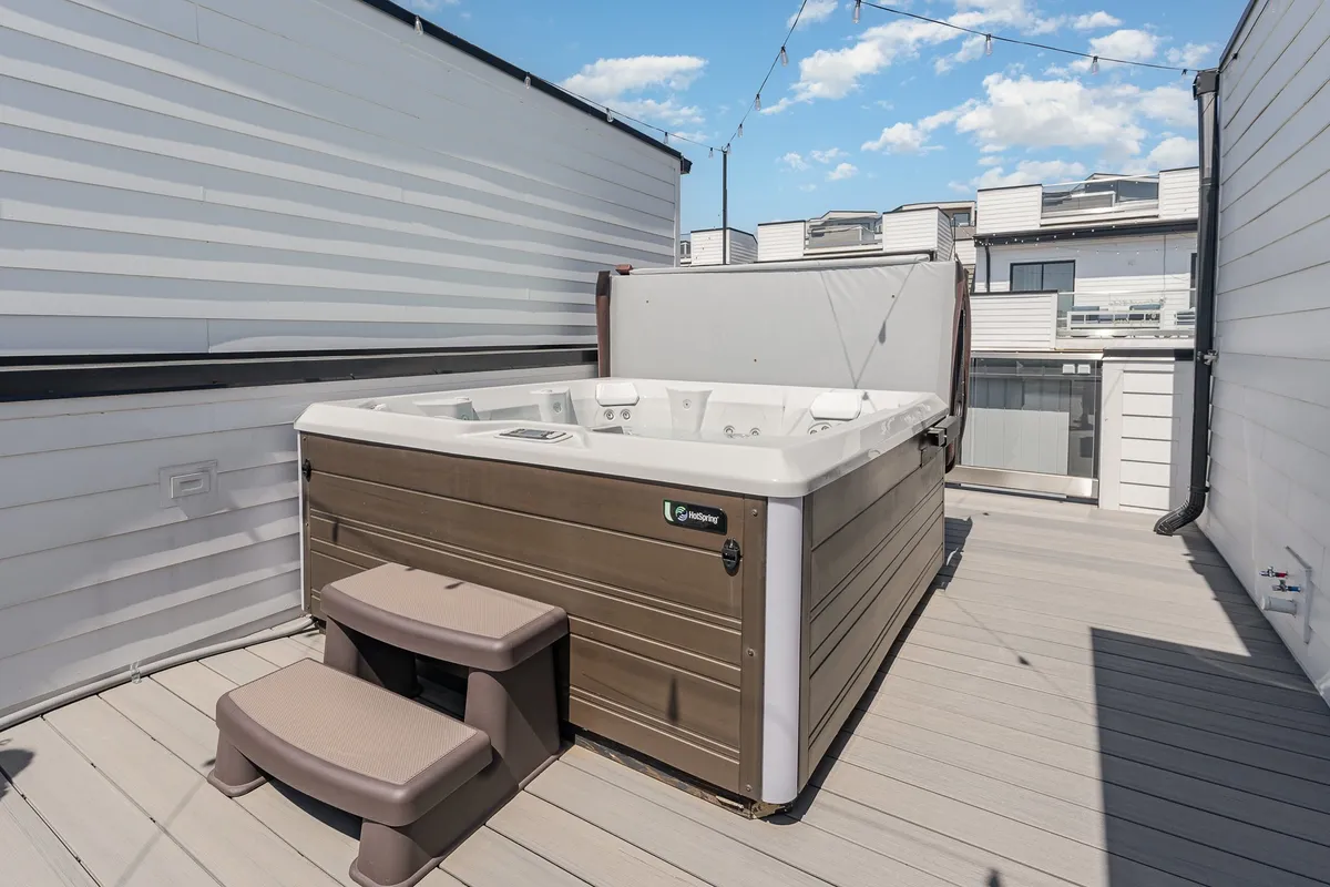 The image features a stylish rooftop deck with a private hot tub as the central focal point, surrounded by wooden decking and illuminated by string lights overhead. The scene exudes a tranquil yet inviting ambiance, with the hot tub offering a luxurious spot to unwind while taking in the views. The deck area is spacious enough for gatherings and relaxation under the open sky.