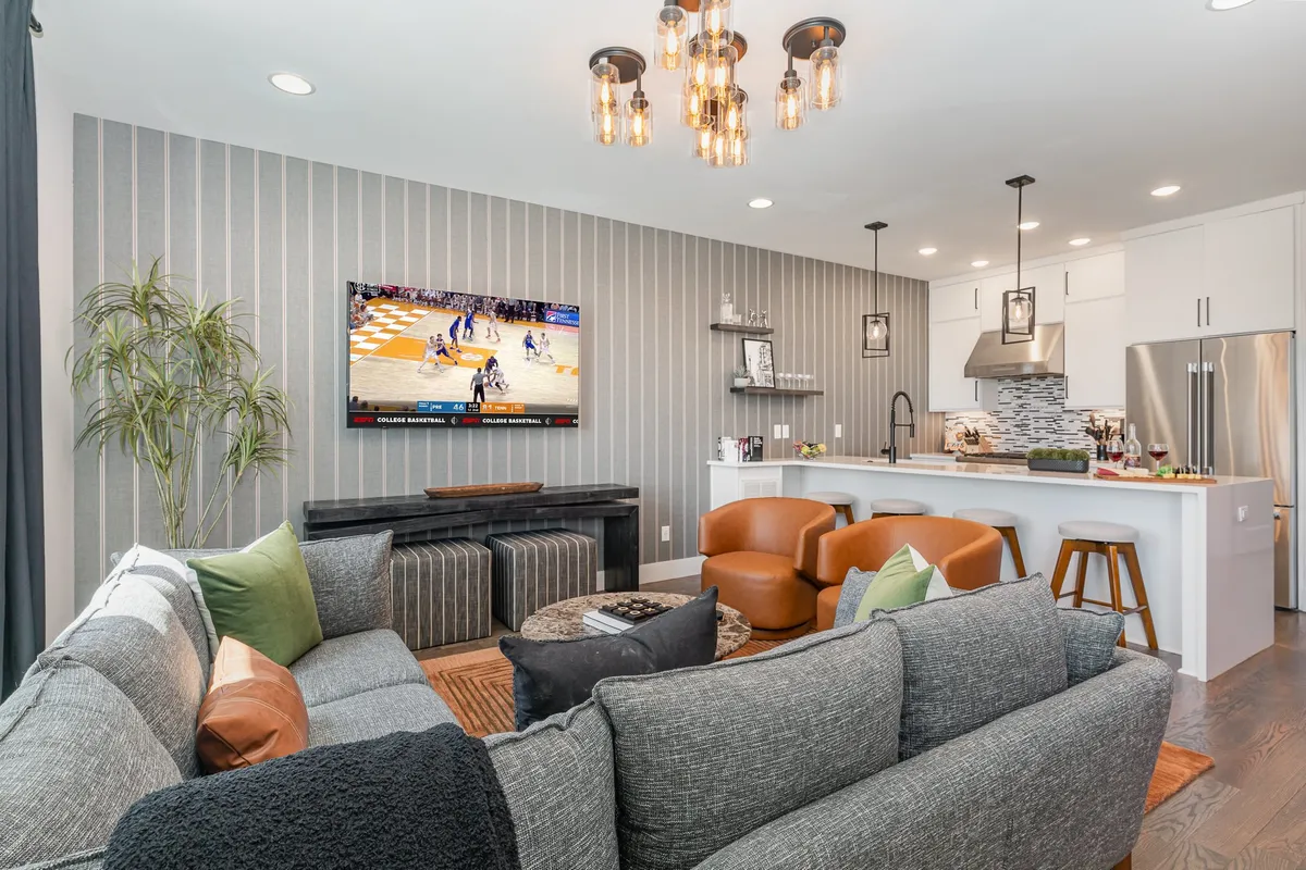 A vibrant living area with a plush sofa adorned with colorful cushions faces a large TV showing a sports game. Nearby, a spacious kitchen island with wooden bar stools invites conversation, while a striking chandelier with exposed bulbs creates a warm, inviting glow. The space hints at lively gatherings, cozy relaxation, and stylish entertaining, all set within a modern, stylish Nashville home.