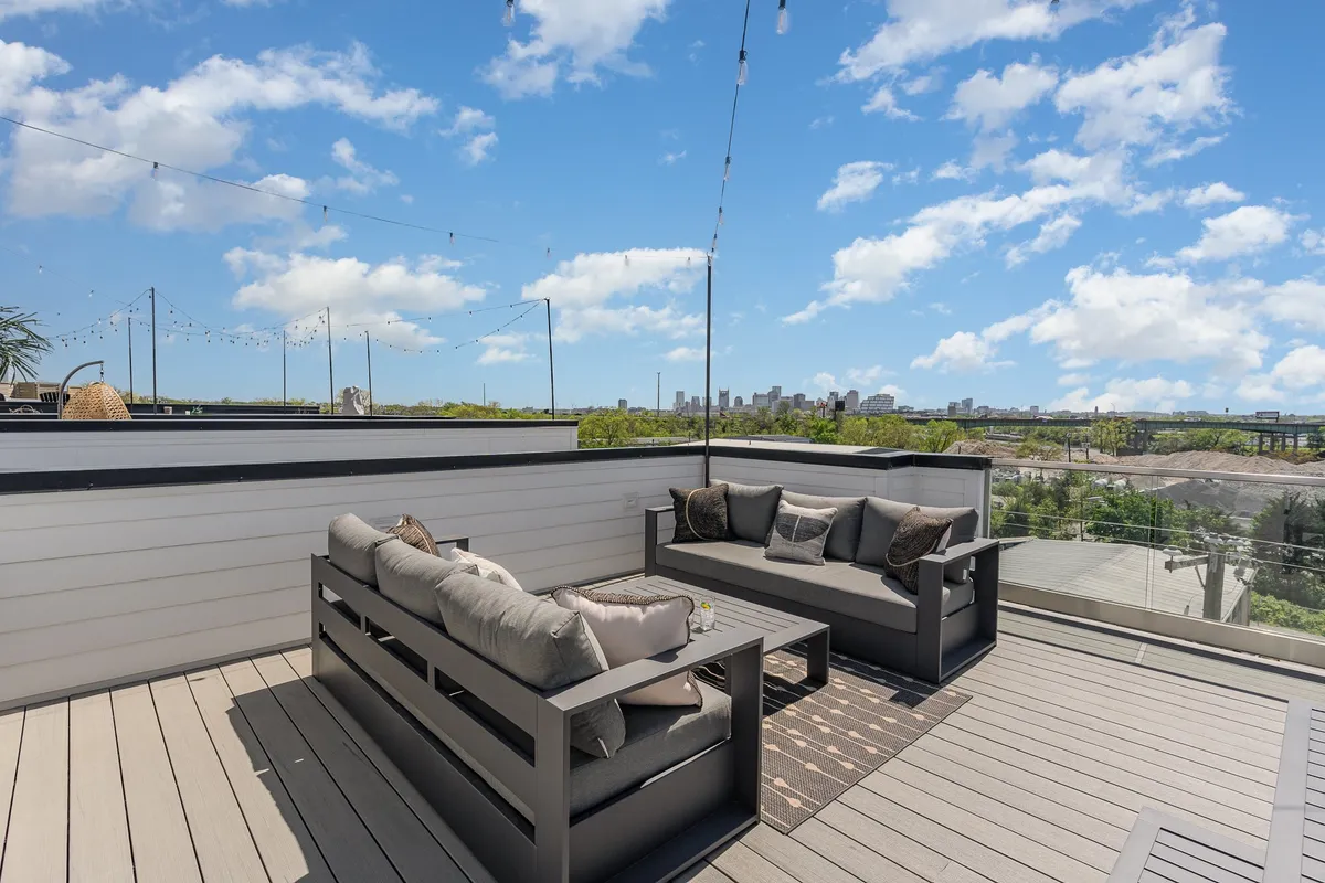 A chic rooftop patio setup featuring modern outdoor lounge seating, a coffee table, and decorative string lights. The city skyline is visible in the background, emphasizing the panoramic urban views. The overall mood is relaxed and stylish, ideal for both social gatherings and quiet moments.