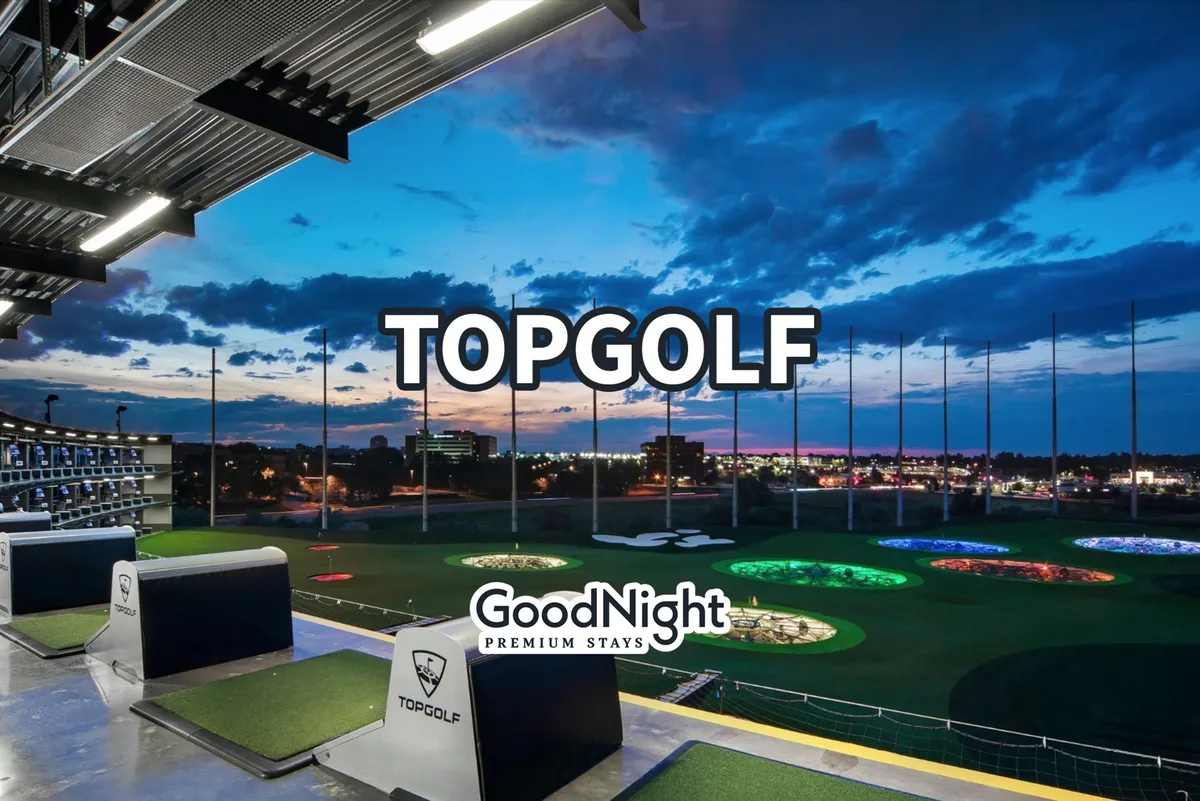 This image features the lively atmosphere of Topgolf, with illuminated golf bays, targets glowing at dusk, and the city skyline visible in the background. Groups of people are seen socializing and enjoying entertainment in a dynamic outdoor setting.