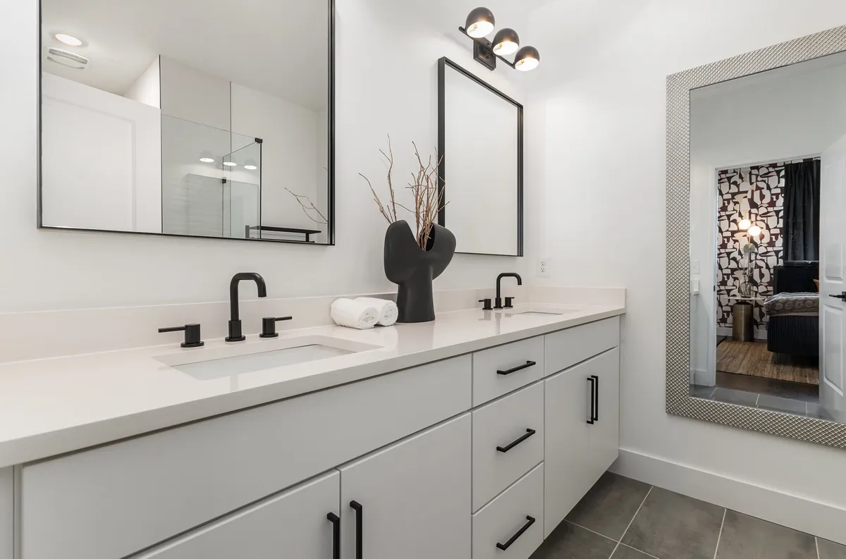 A pristine bathroom featuring dual mirrors above a chic vanity, complemented by black faucet fixtures and a decorative vase with branches. The overall aesthetic is clean and contemporary, with a focus on tranquility and modern comfort.